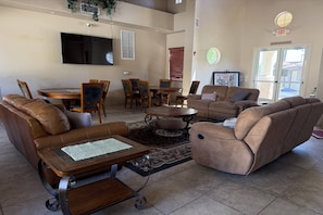 A spacious living room with a large flat-screen TV, leather sofas, a dining table with chairs, and a large window with blinds. - 3BR Mesa Getaway | Pool, Hot Tub, Gym, Clubhouse, Basketball + Room for the Whole Family (Mesa)