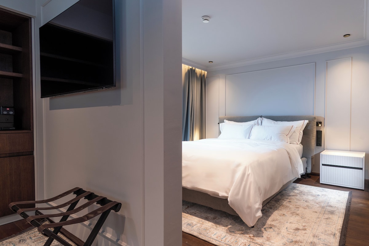 A modern bedroom with a large bed, a TV mounted on the wall, a bedside table, and a wooden bench.