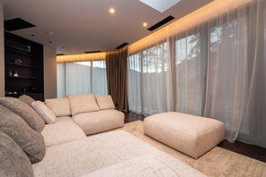 Una sala moderna con un sofá modular, un reposapiés redondo y cortinas traslúcidas.