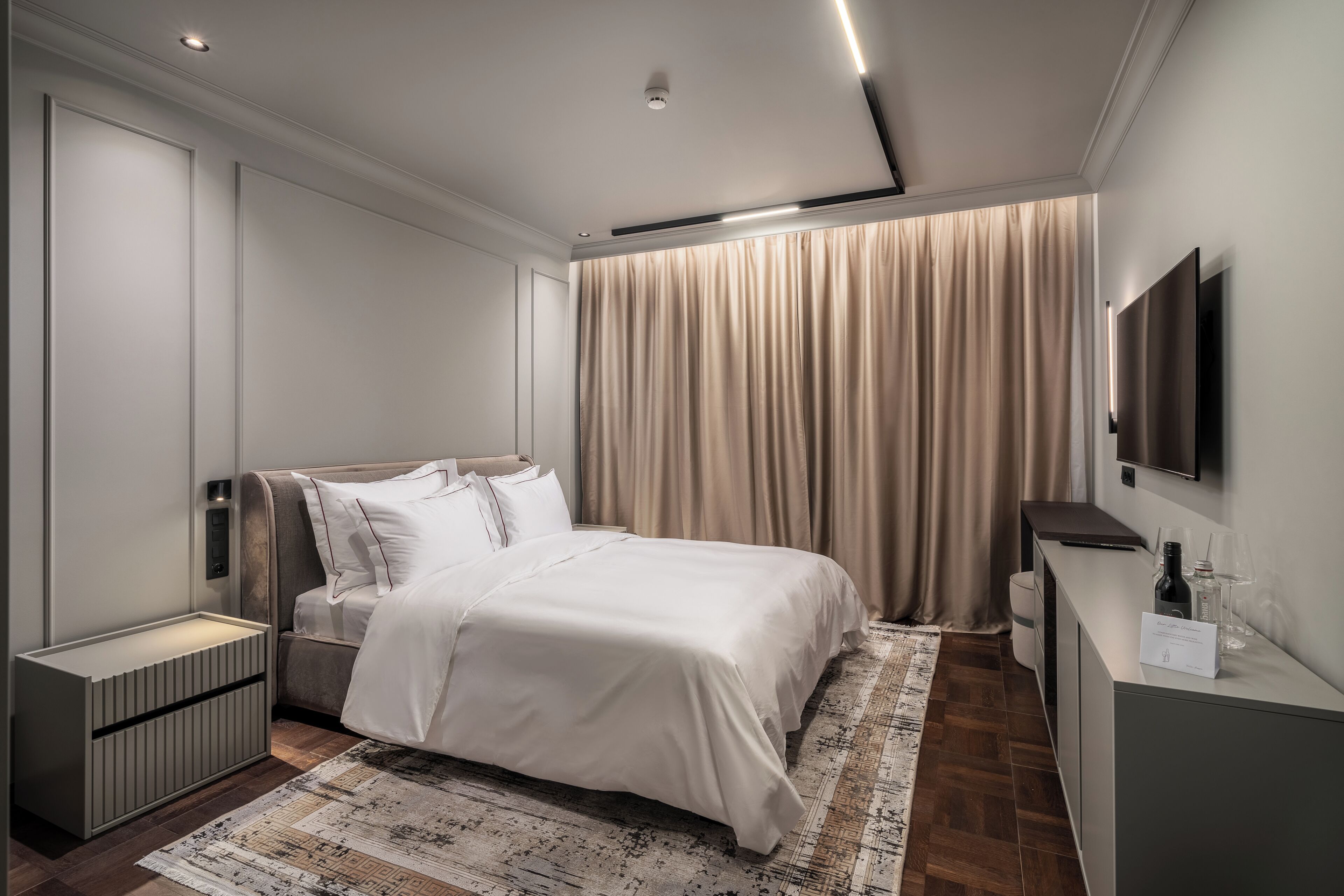 A modern hotel bedroom with a large bed, bedside tables, a flat-screen TV, and a patterned rug.
