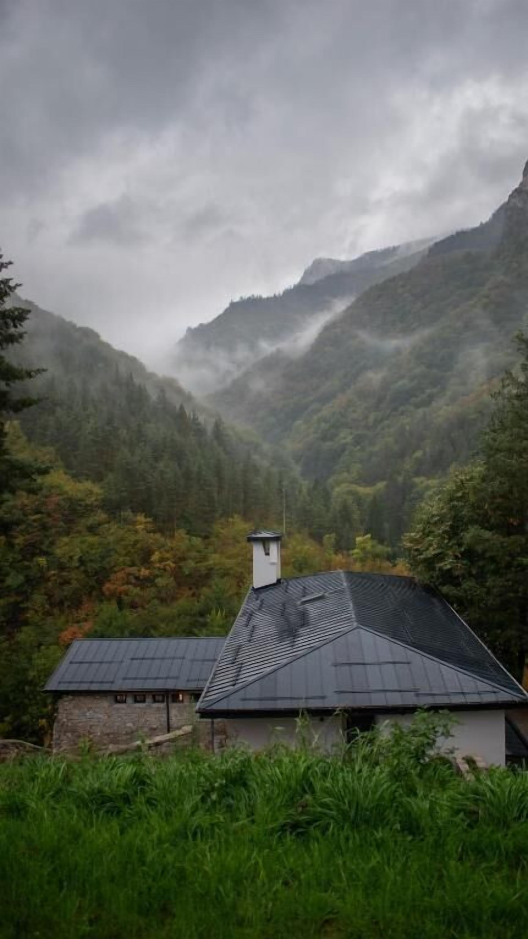 A house with a sloped roof, surrounded by dense forest and mountains.