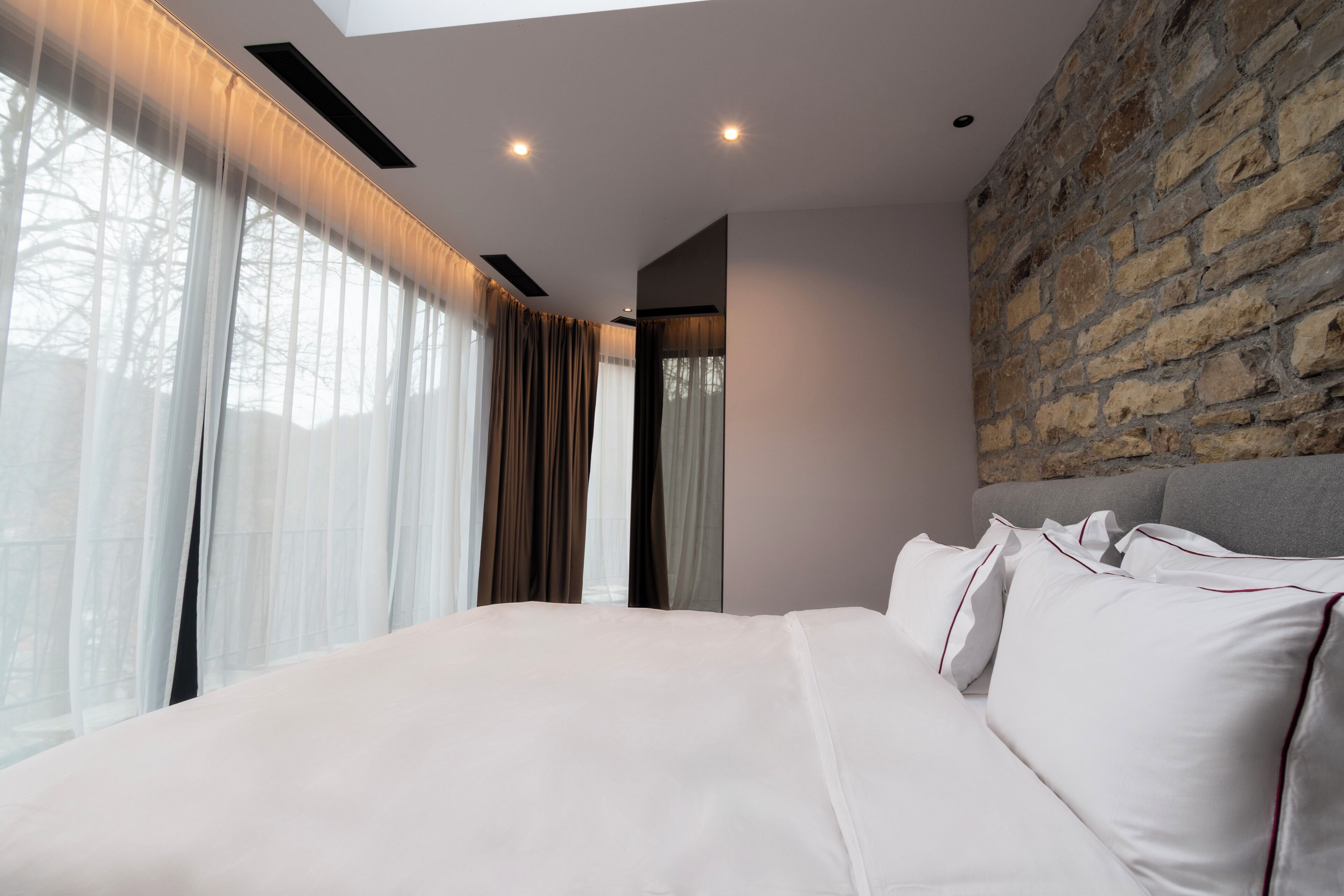 A modern bedroom with a large bed, a stone accent wall, and sheer curtains.