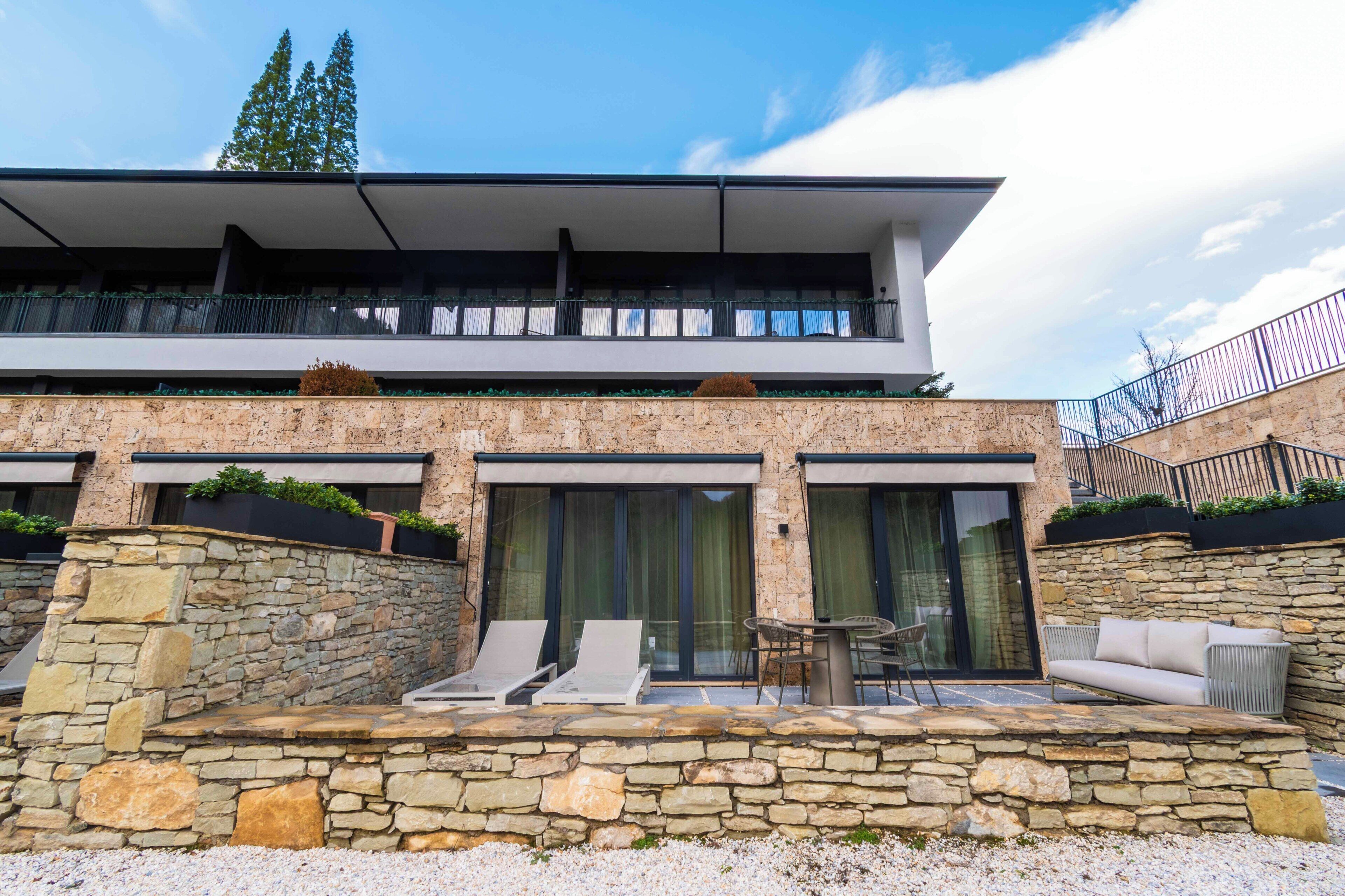 A modern two-story building with a stone facade, a balcony with a railing, and a seating area with a table and chairs.