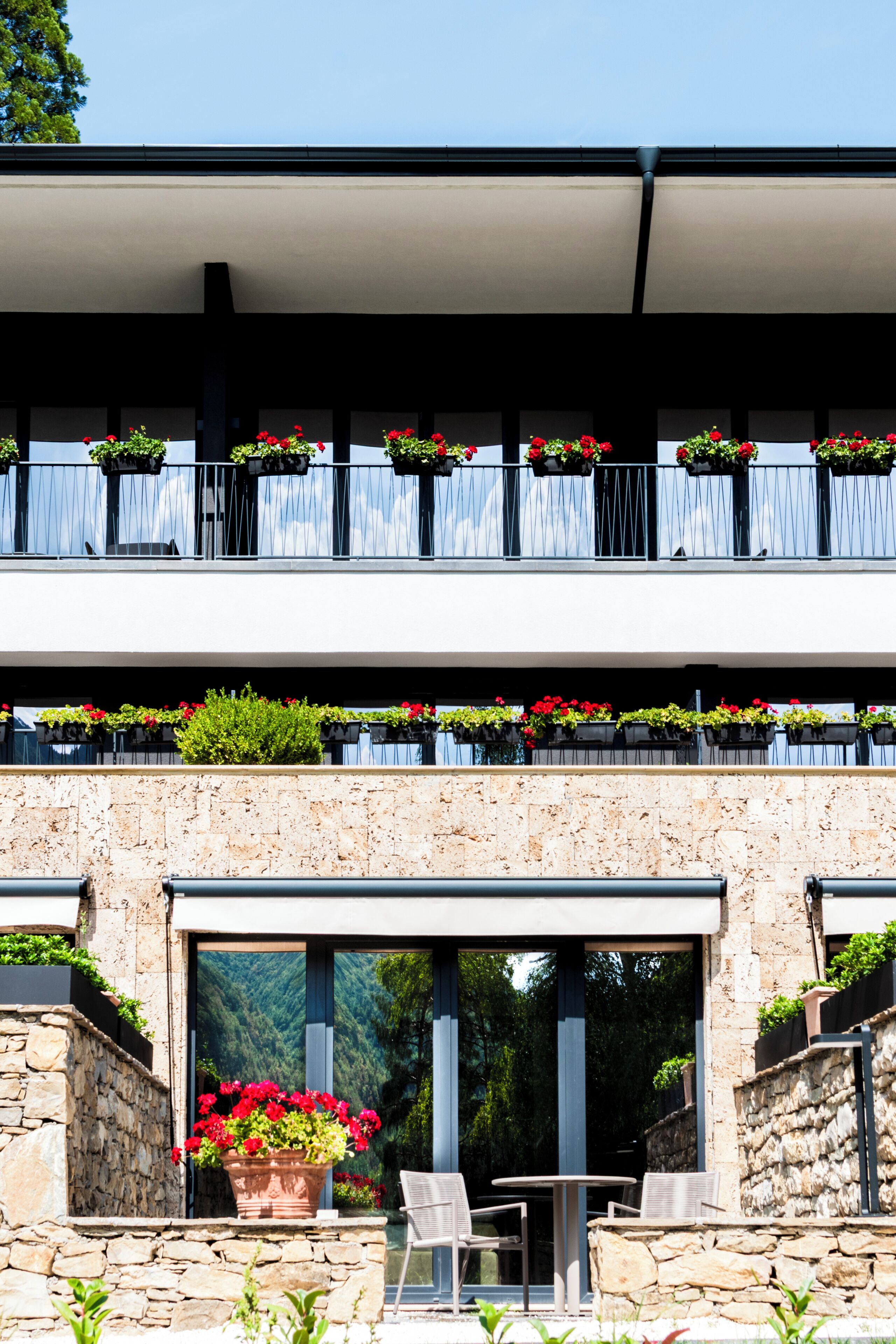 Un edificio moderno con fachada de piedra, un balcón con jardineras de flores y una puerta de vidrio que da a una zona de estar al aire libre.