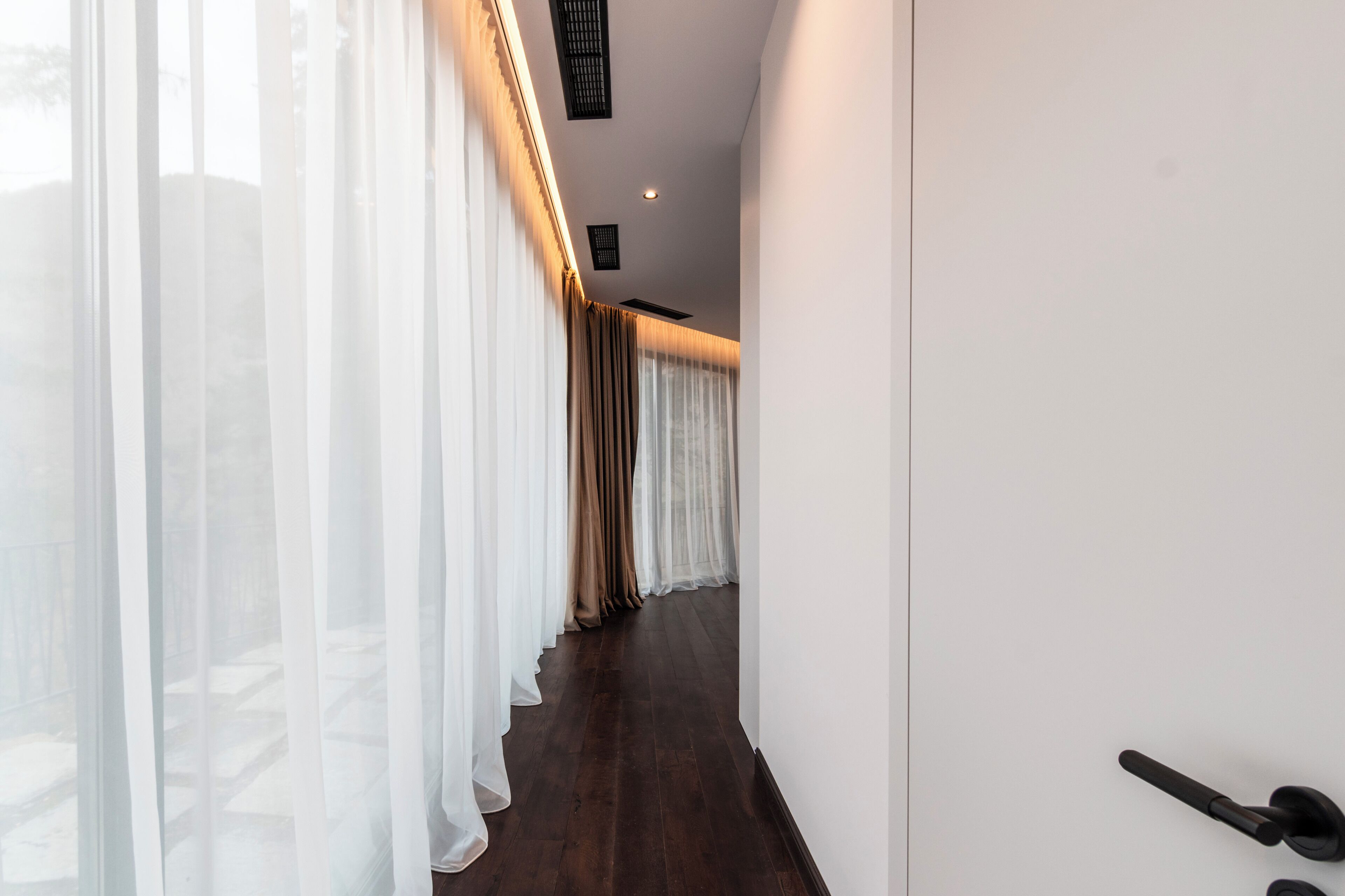 Un pasillo con varias cortinas blancas, suelo de madera y una puerta con pomo negro.