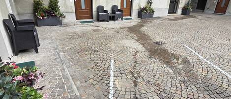 A courtyard with a cobblestone floor, outdoor seating, and a building with multiple doors and windows.