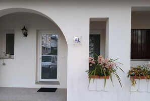 Um edifício branco com uma porta, uma placa com a letra 'AL' e vasos de flores à direita.