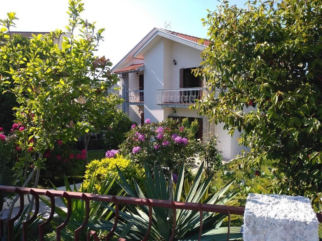 A two-story building with a balcony, surrounded by lush greenery and flowering plants.