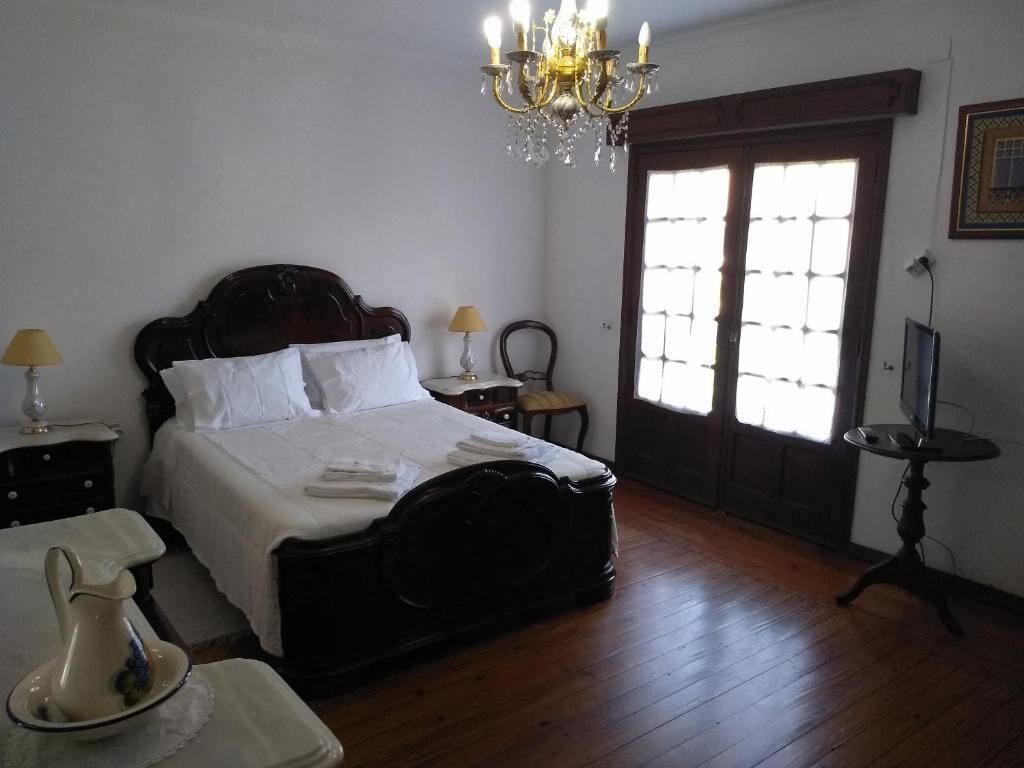 Une chambre à coucher avec un lit en bois, un lustre, une fenêtre avec des stores et un téléviseur posé sur un meuble.