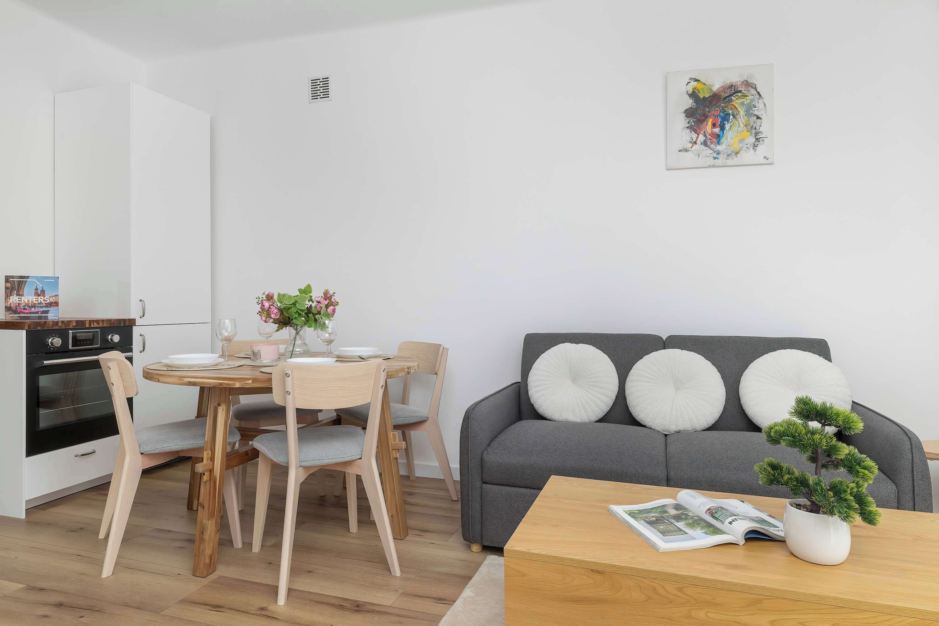 A modern living space with a dining area, a grey sofa with white cushions, and a small coffee table with a magazine and a plant.