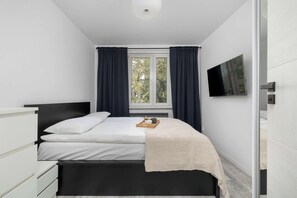 Ein Schlafzimmer mit einem Bett, einem Nachttisch, einem Fenster mit Vorhängen, einer Heizung und einem an der Wand befestigten Fernseher.