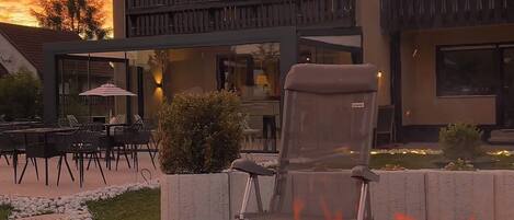 A fire pit with flames in front of a building with a balcony and outdoor seating.