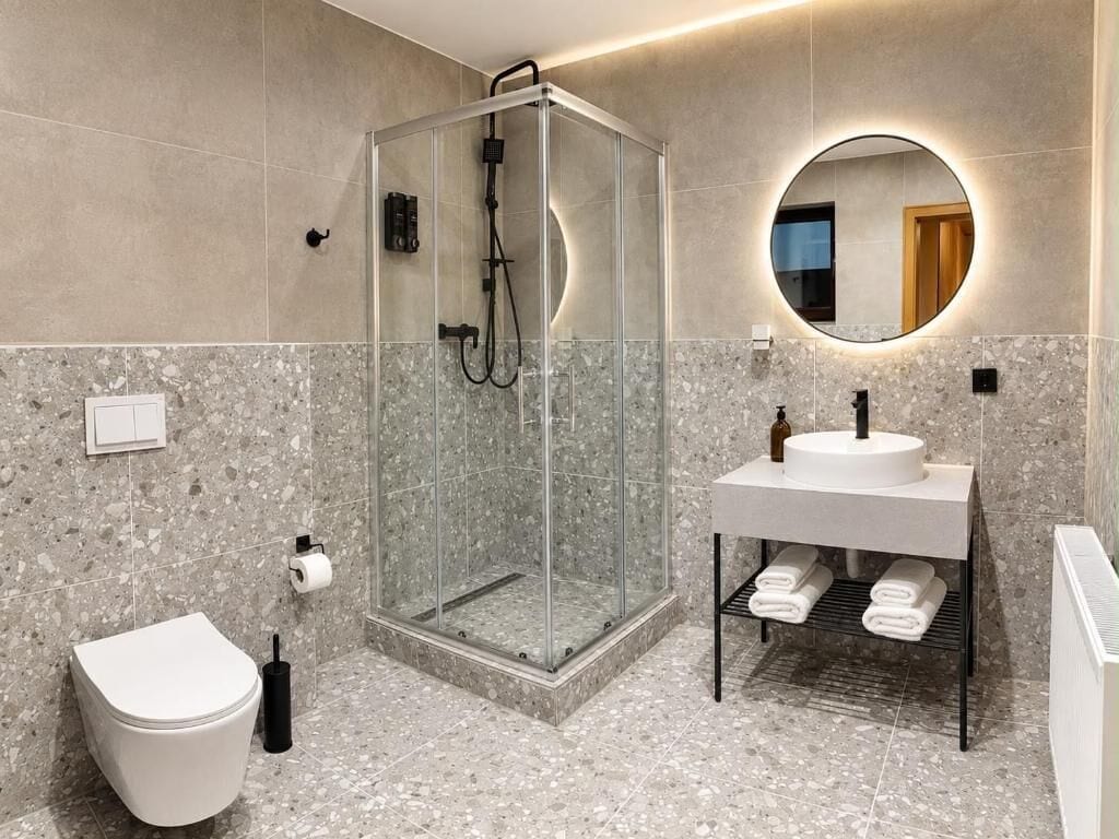 Et moderne badeværelse med en glasafskærmet brusekabine, et hvidt toilet, en vask med håndklædestang og et rundt spejl.