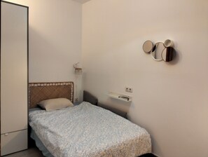 Une petite chambre avec un lit simple, un mur uni, une table de chevet et un miroir fixé au mur.