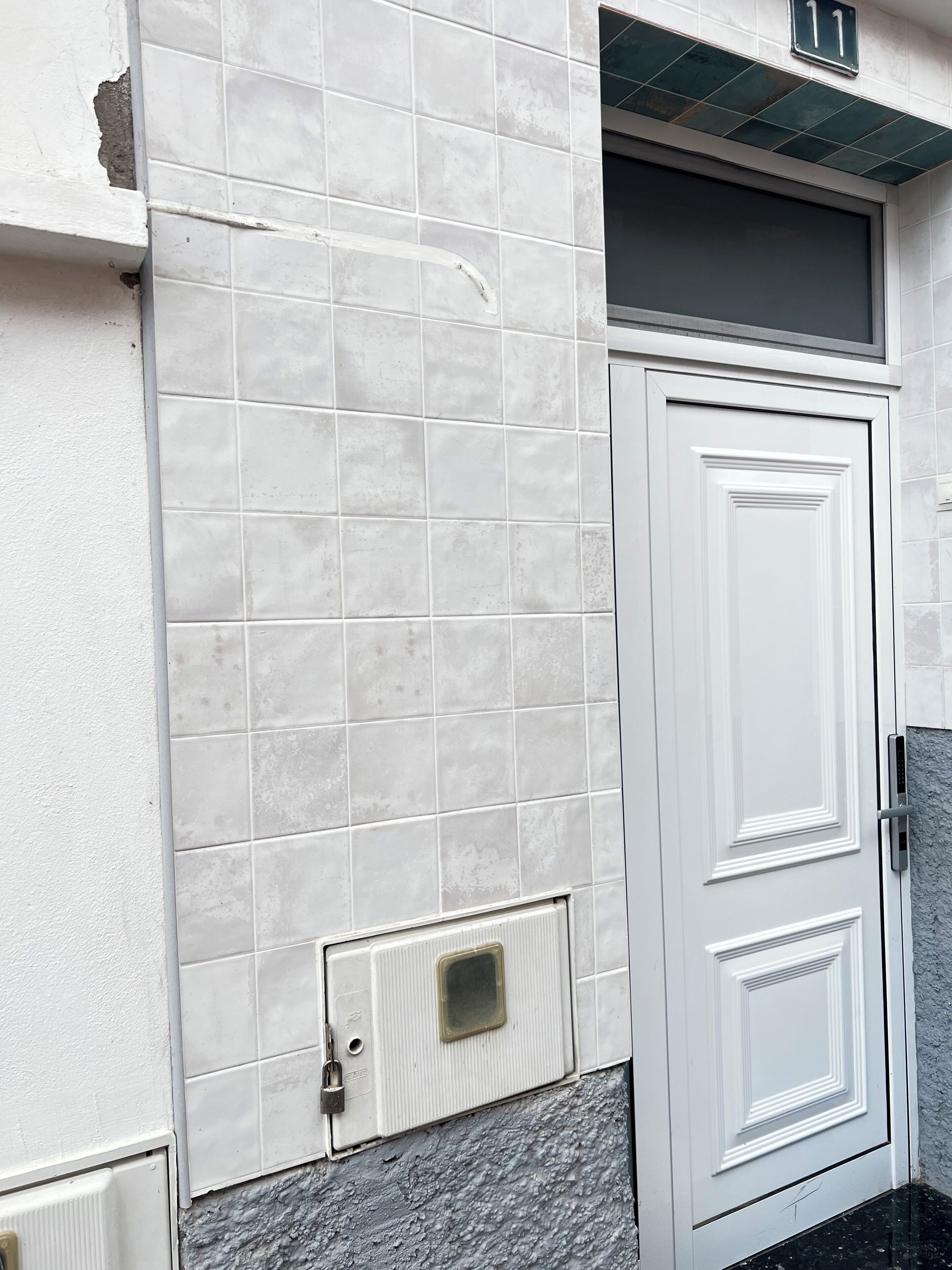 Une porte blanche dotée d’un clavier pour ouvrir, une grille d’aération en dessous et un mur recouvert de carreaux, avec un panneau indiquant un numéro au-dessus.
