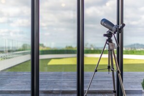 A telescope on a tripod inside a room with large windows overlooking a green field. - Luxury 3BR Apartment with Stunning Views | Remuera (Auckland)