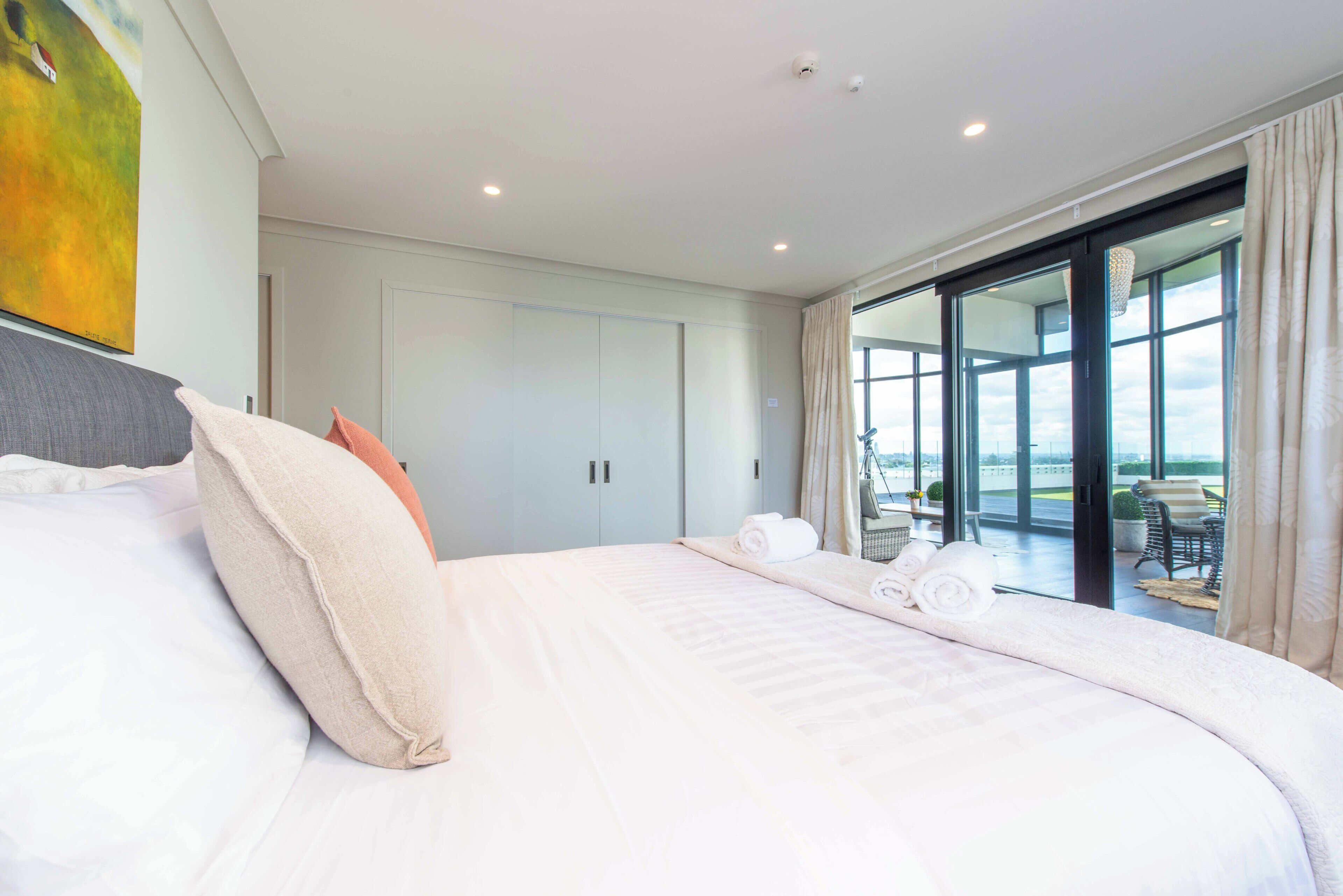 A modern hotel room with a large bed, a view of the outdoors through a sliding glass door, and a painting on the wall.