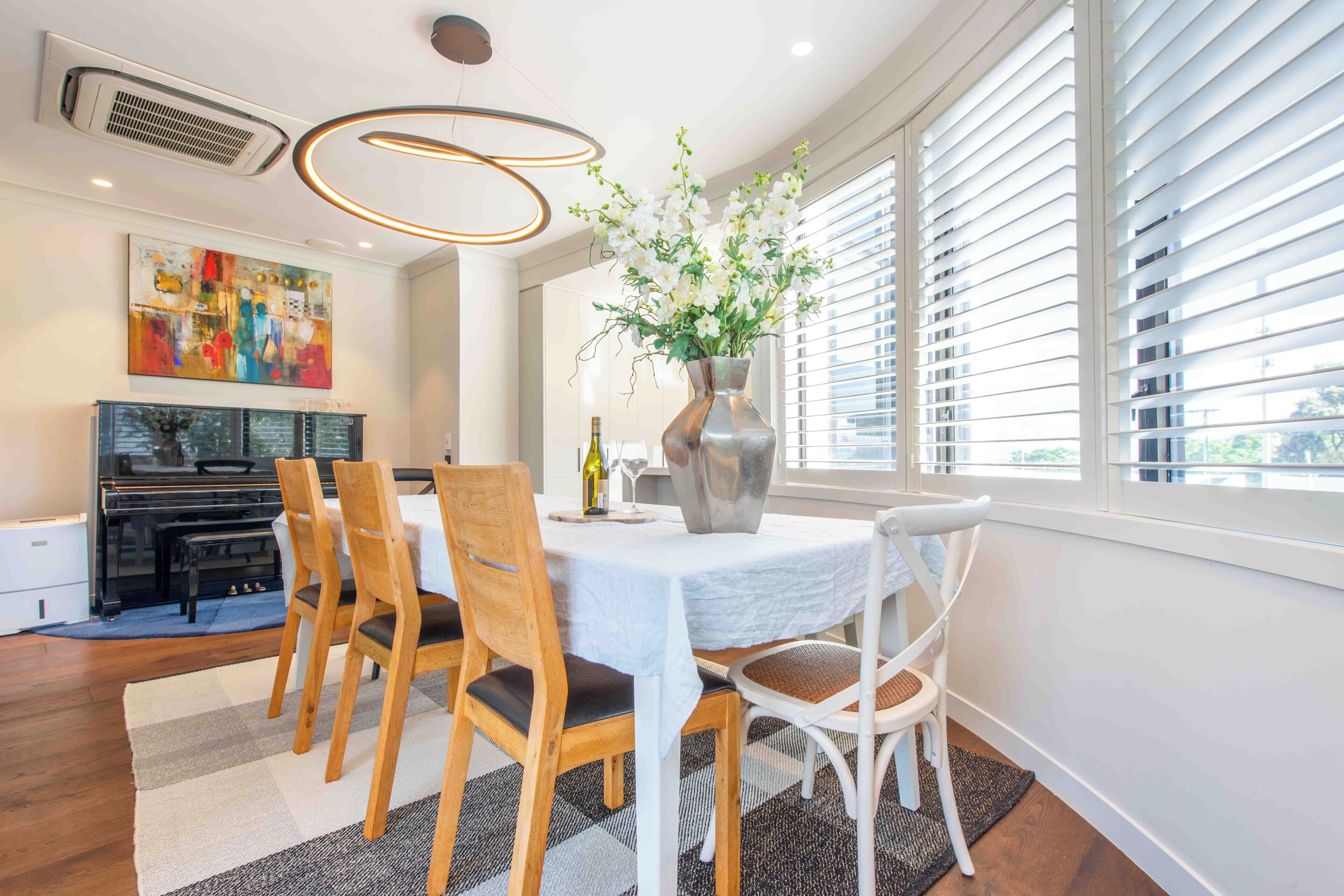 A dining area with a piano, a table with chairs, a vase with flowers, and a large window with blinds.