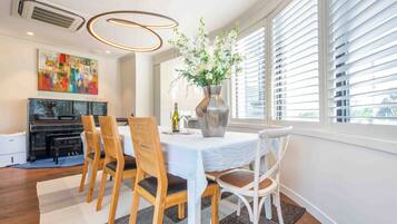 A dining area with a piano, a table with chairs, a vase with flowers, and a large window with blinds.