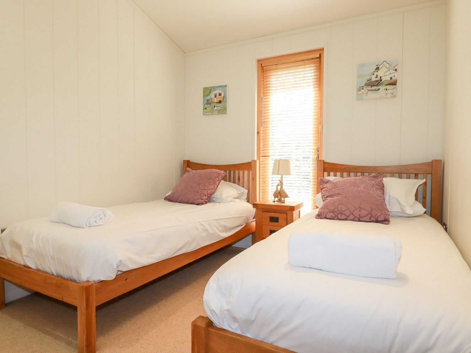 Two single beds with wooden frames and white bedding, a nightstand with a lamp, and a window with blinds.