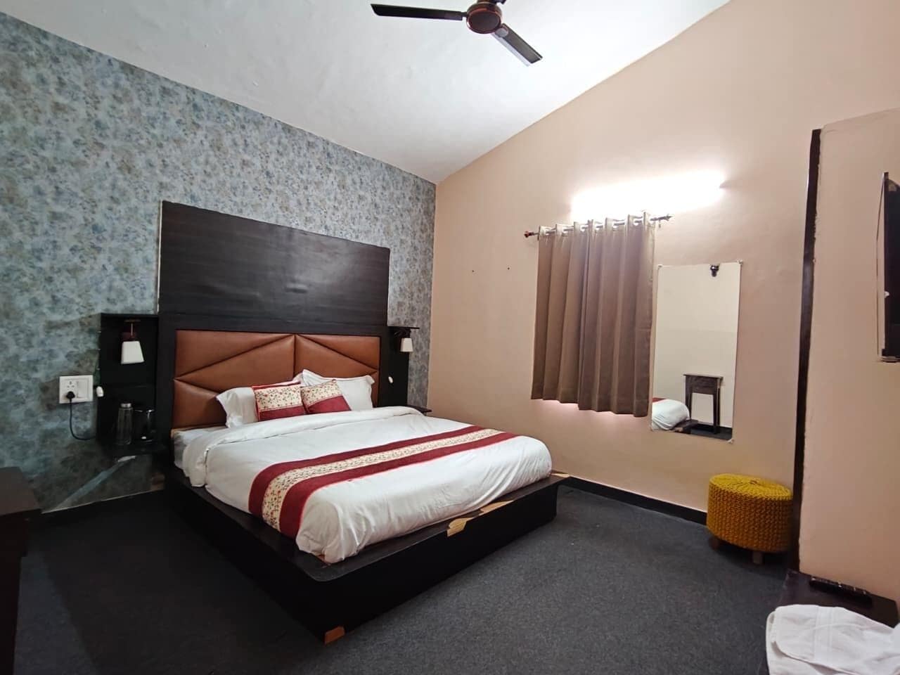 Ein Hotelzimmer mit einem Bett, einem Deckenventilator, einem Nachttisch, einer Lampe, einem Spiegel und einem kleinen Hocker.
