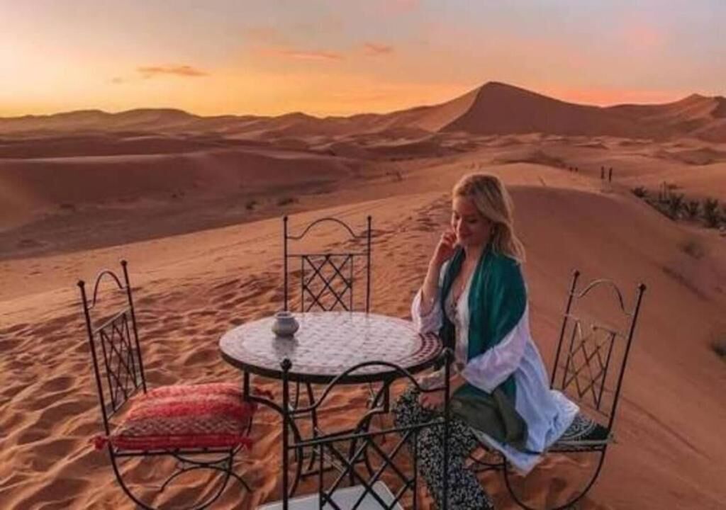 A desert scene with a person sitting at a round table with chairs, surrounded by sand dunes and a mountain in the background.