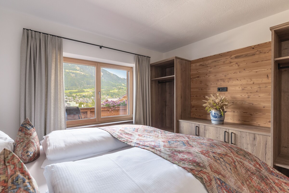 A bedroom with a large window, a bed with white linens, a wooden wardrobe, and a potted plant.