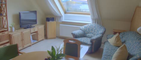 Un salon comprenant une télévision, un canapé, un fauteuil, une table avec une plante et une fenêtre avec des rideaux.