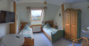 Ein Schlafzimmer mit zwei Einzelbetten, einem Holzschrank, einem Nachttisch, einem kleinen runden Tisch, einem Stuhl, einem an der Wand montierten Fernseher und einem Fenster mit Vorhängen.