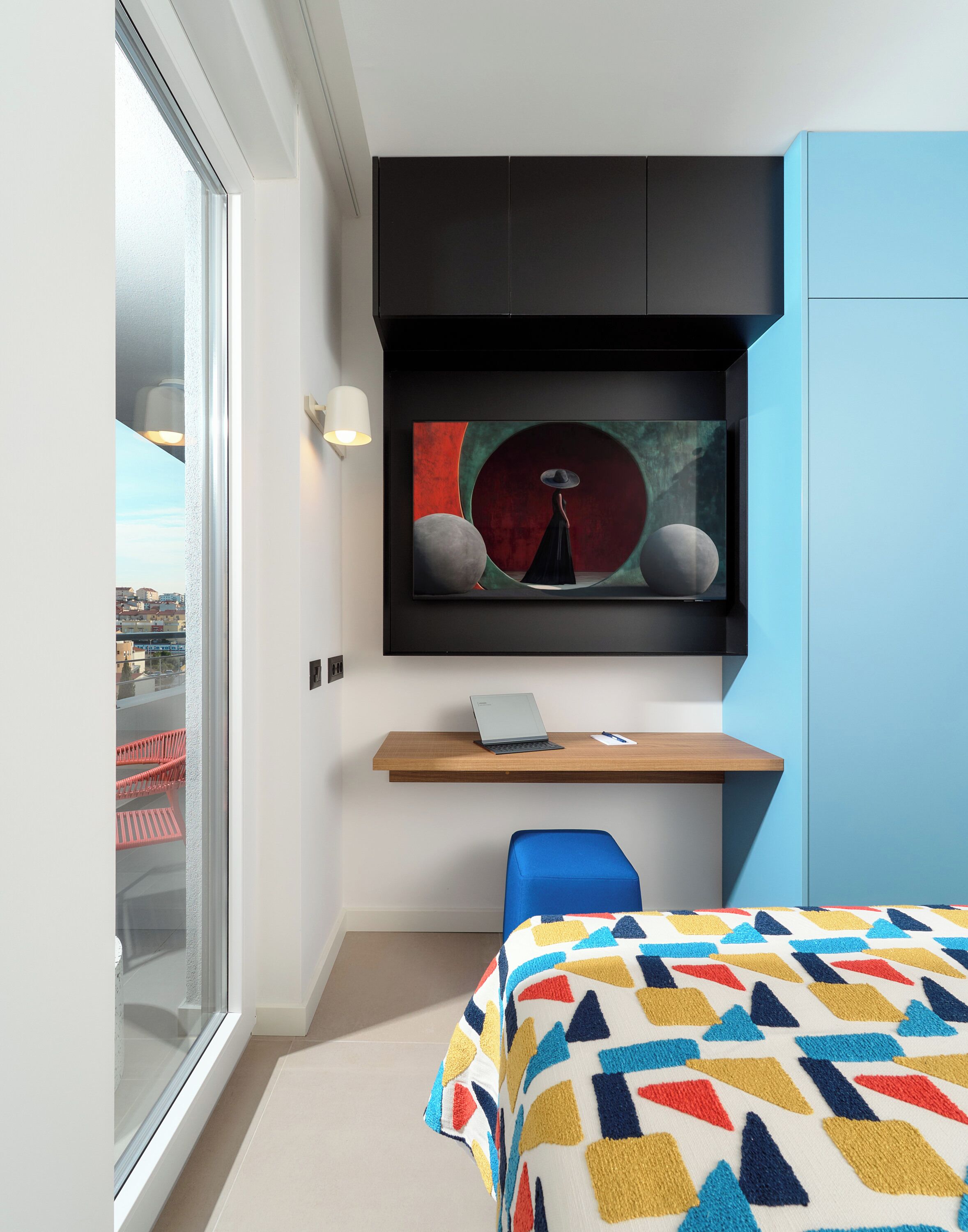 A modern bedroom with a large window, a bed with a colorful geometric-patterned bedspread, a desk with a laptop, and a wall-mounted artwork.