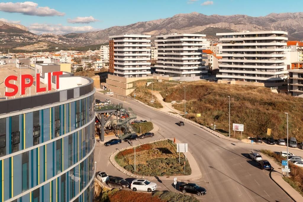 A modern building with a curved facade and multiple floors, surrounded by a parking area and a road with vehicles.