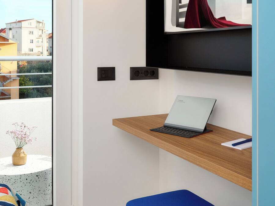 Ein modernes Zimmer mit einem Laptop auf einem Holzbodenregal, einem blauen Ottomanen und einer Wandlampe.