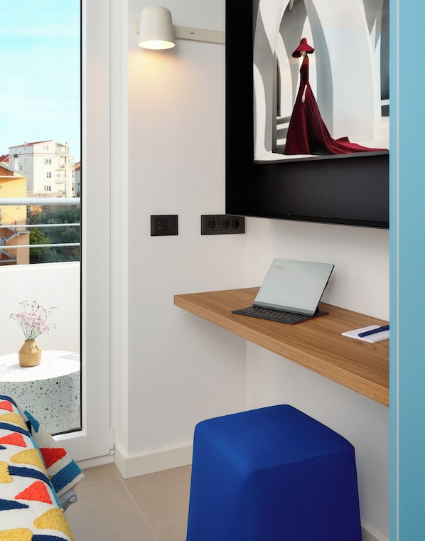 Una habitación moderna con una computadora portátil sobre un estante de madera, un otoman azul y una lámpara fijada a la pared.
