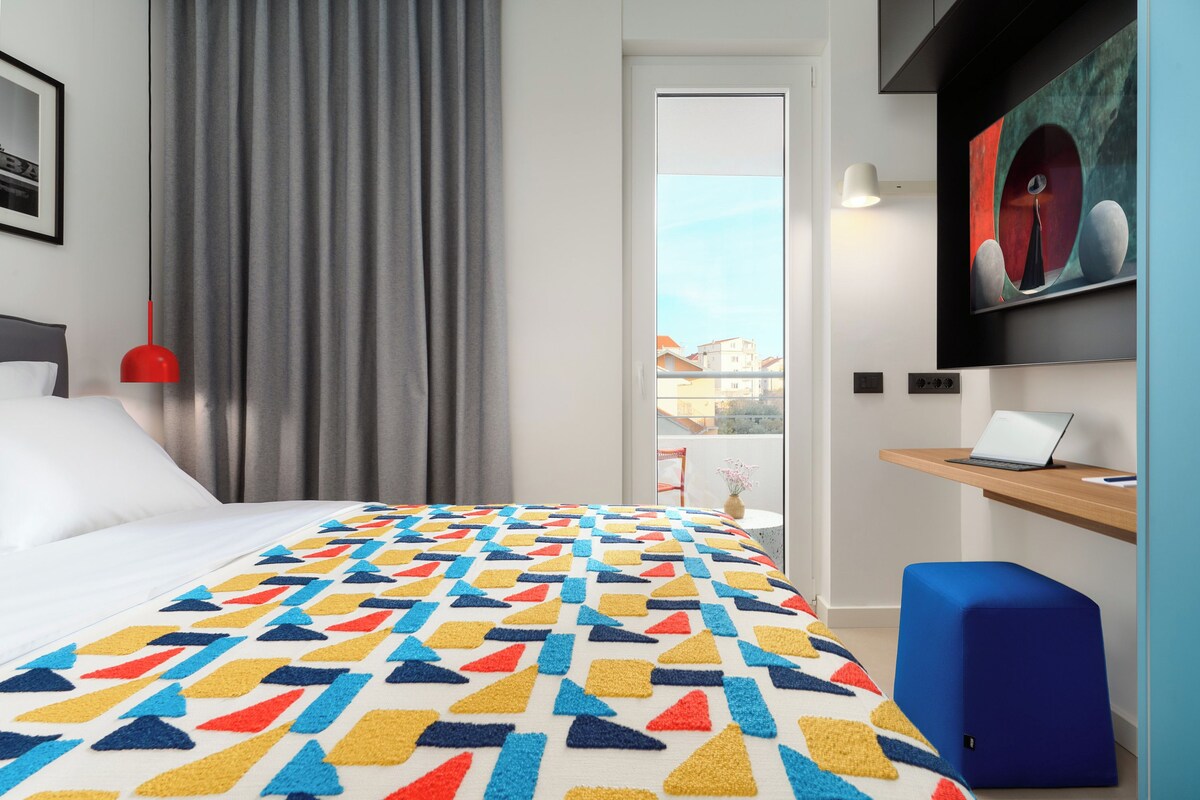 A hotel room with a bed, a desk with a laptop, a chair, a window with a view of buildings, and a painting on the wall.