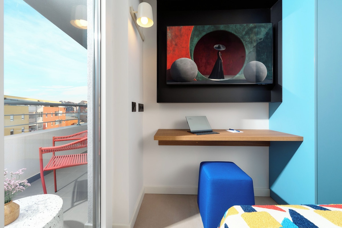 Una habitación moderna con escritorio, una computadora portátil, una silla azul, un sofá y un cuadro en la pared.