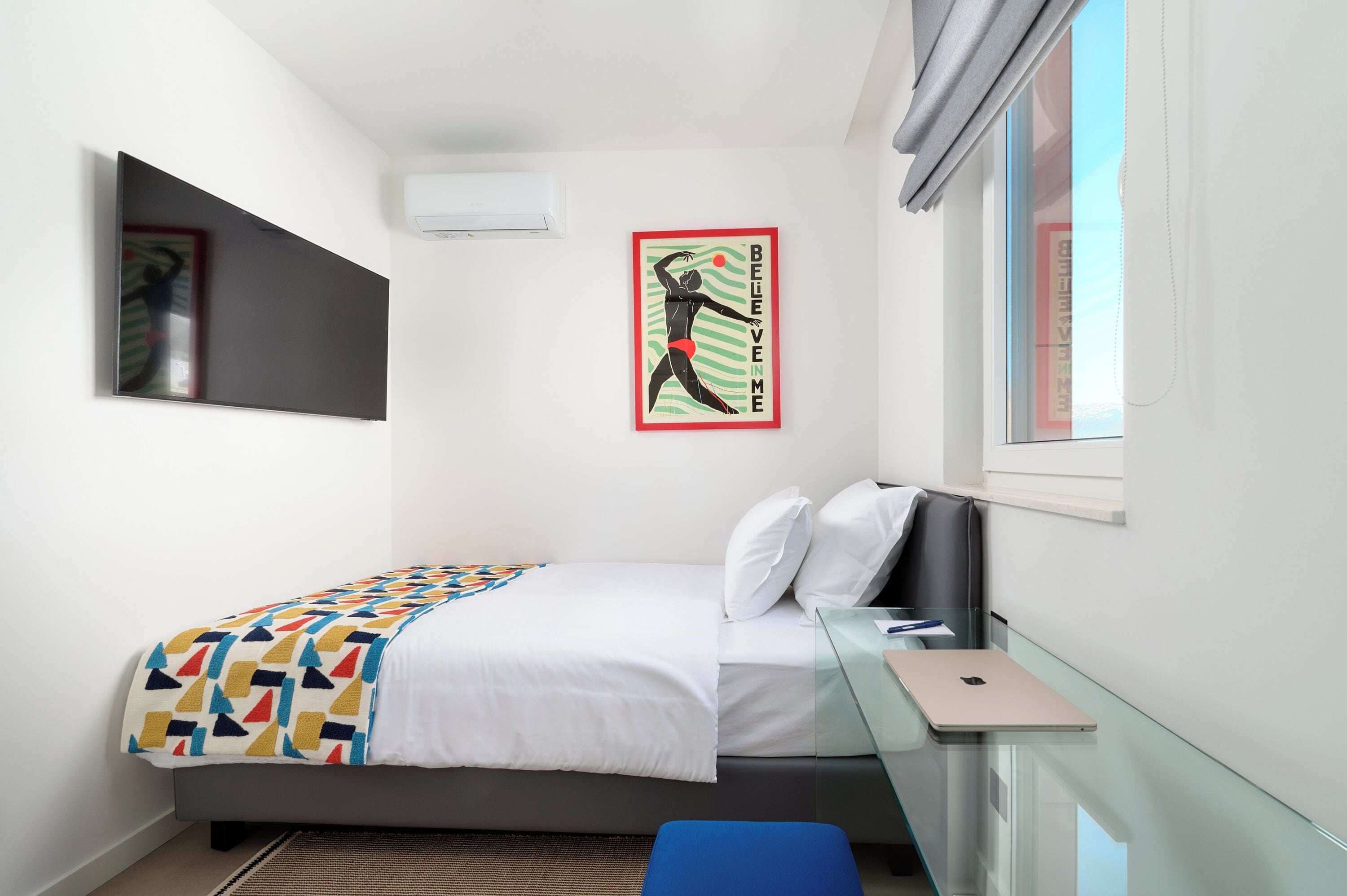 Un dormitorio con cama, escritorio, ordenador portátil, ventana y aire acondicionado instalado en la pared.