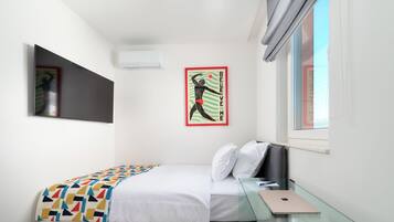 Un dormitorio con cama, escritorio, ordenador portátil, ventana y aire acondicionado instalado en la pared.