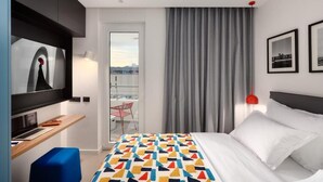 Habitación de hotel con cama, escritorio, laptop, televisor, una silla roja y balcón con vistas.