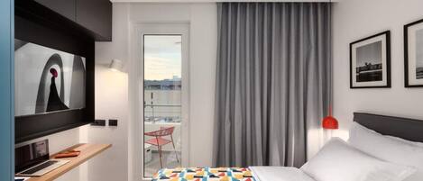A hotel room with a bed, a desk with a laptop, a television, a red chair, and a balcony with a view.