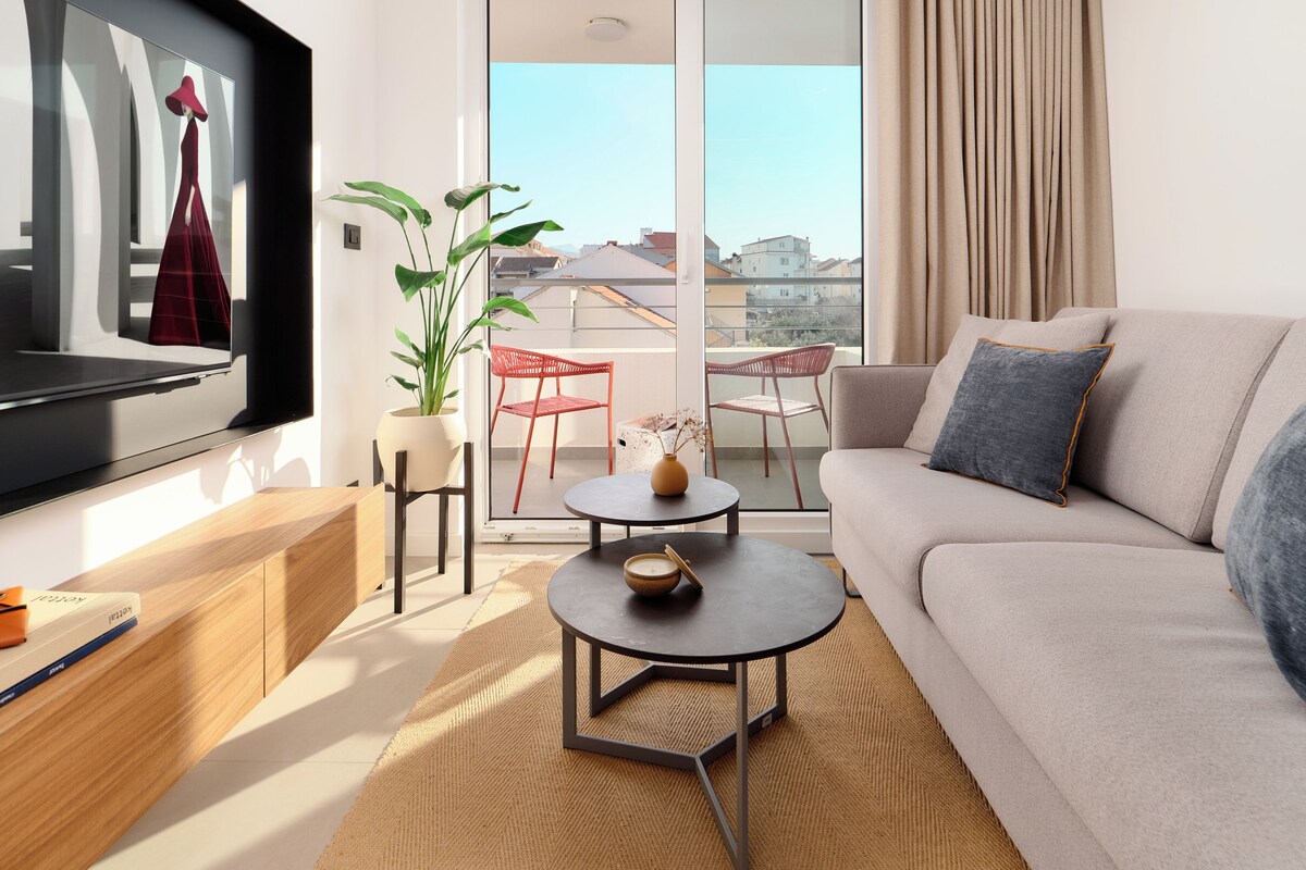A modern living room with a sofa, a coffee table, a TV, and a balcony with plants and chairs.