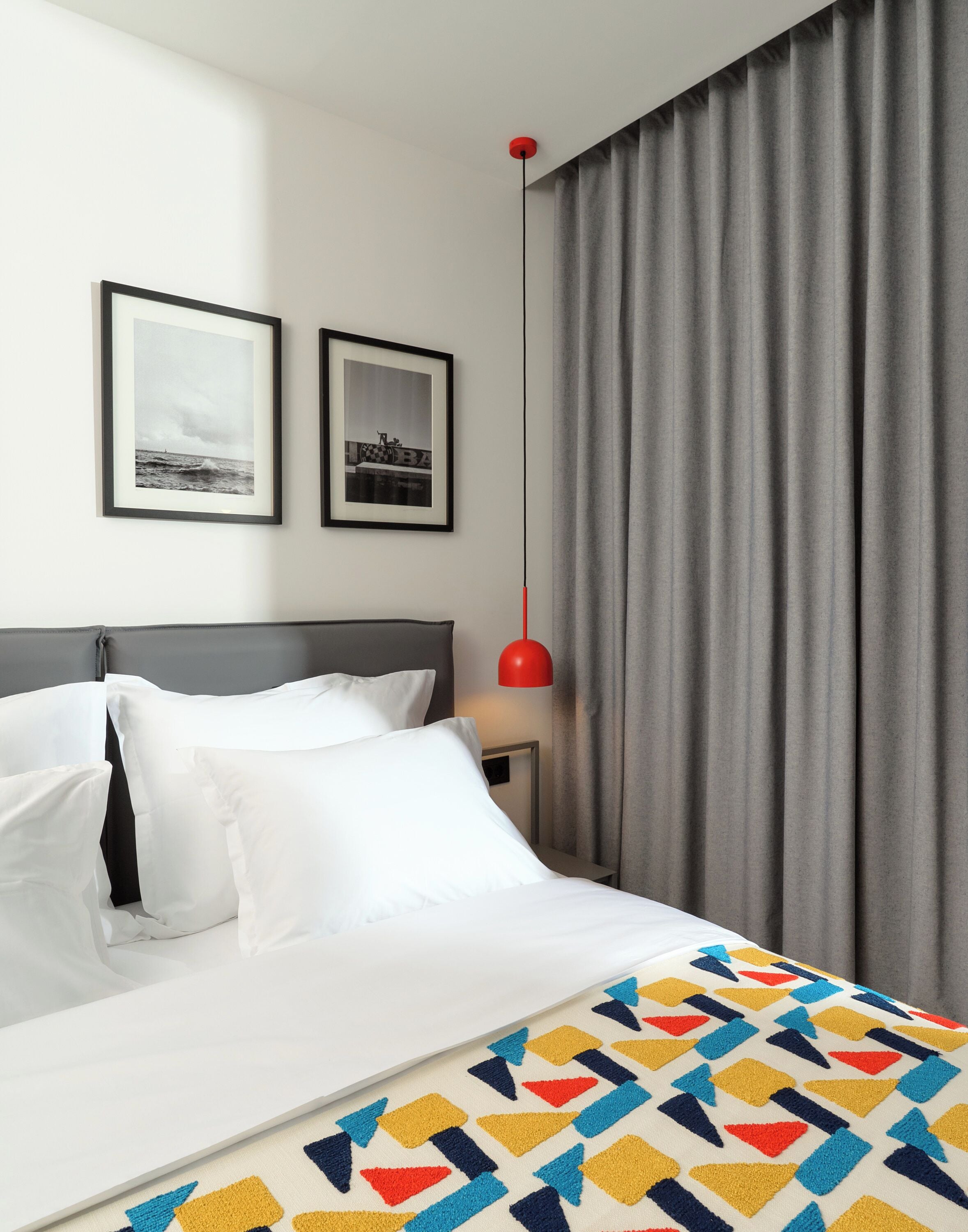 A hotel room with a bed, two framed pictures on the wall, a red pendant light, and grey curtains.