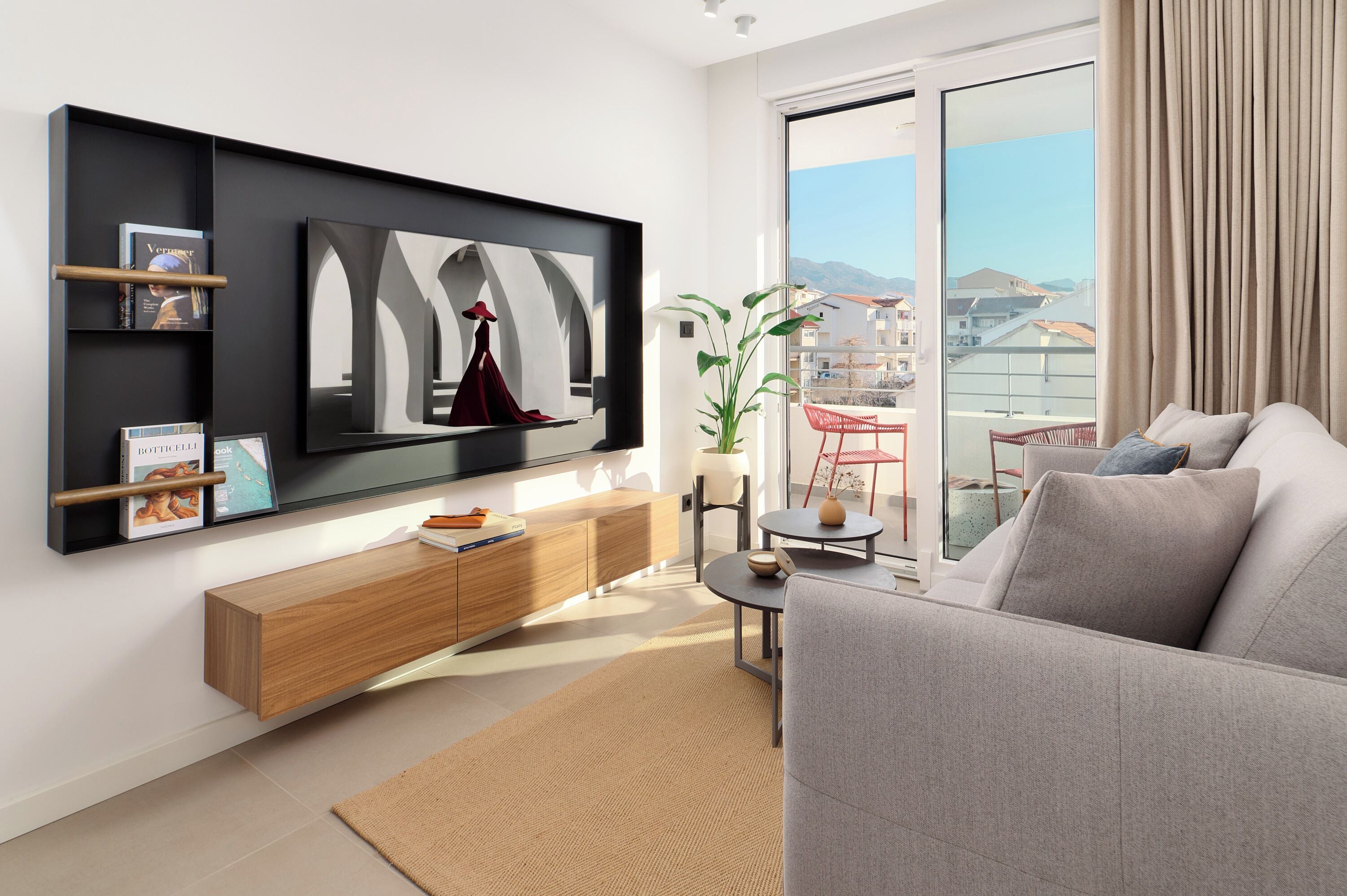 A modern living room with a flat-screen TV mounted on the wall, a grey sofa, a wooden console table, and a view of a balcony with a cityscape.