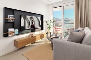 Una sala de estar moderna con un televisor de pantalla plana montado en la pared, un sofá gris, una mesa consola de madera y vistas a un balcón con un paisaje urbano.