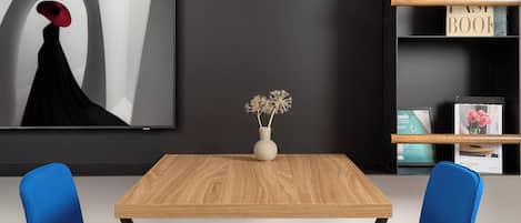 A modern room with a wooden table, two blue chairs, a white lamp, and a black shelf with books.