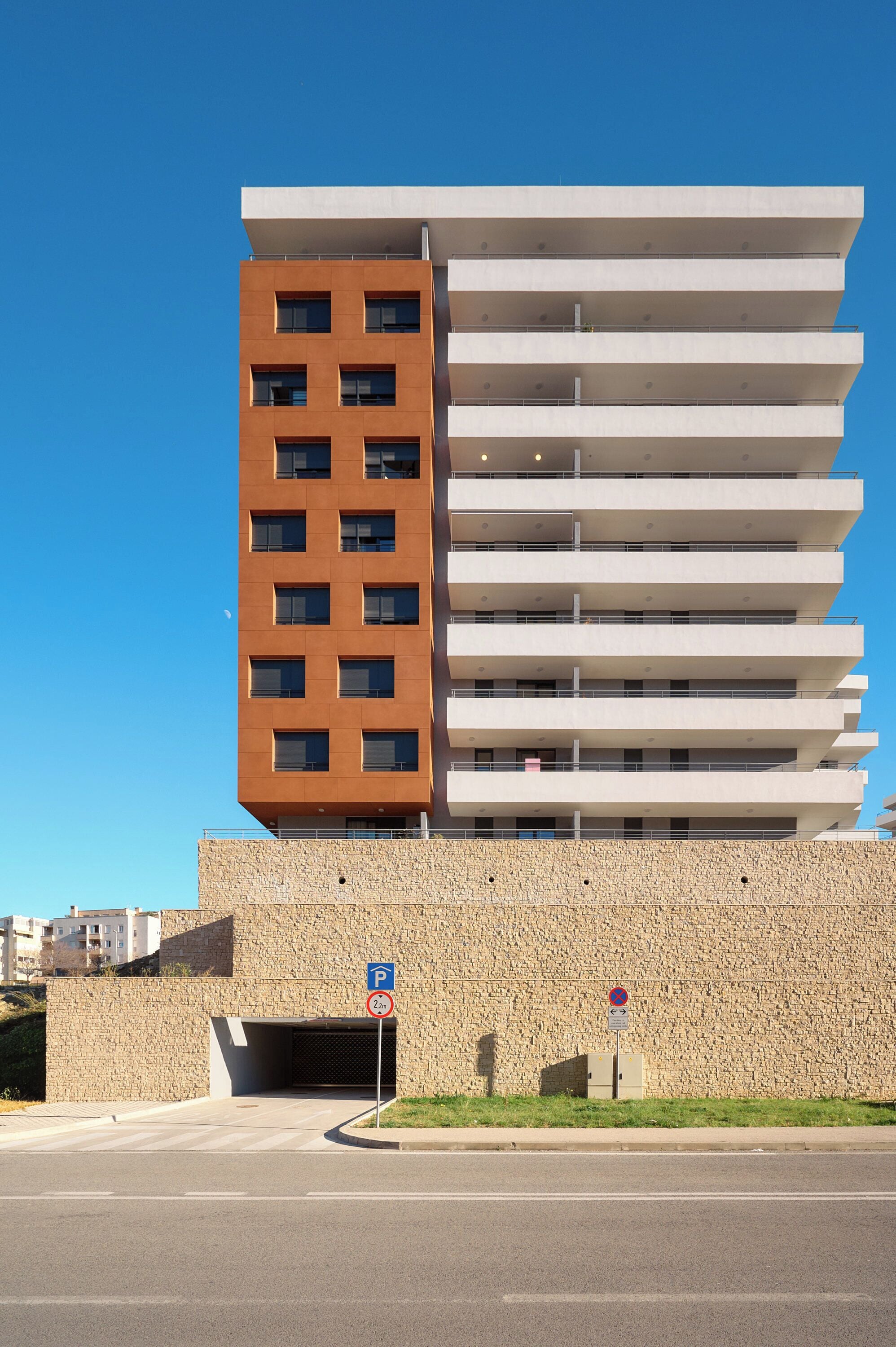 Um edifício moderno de vários andares com fachadas em tons de marrom e branco, entrada de estacionamento e céu azul claro.