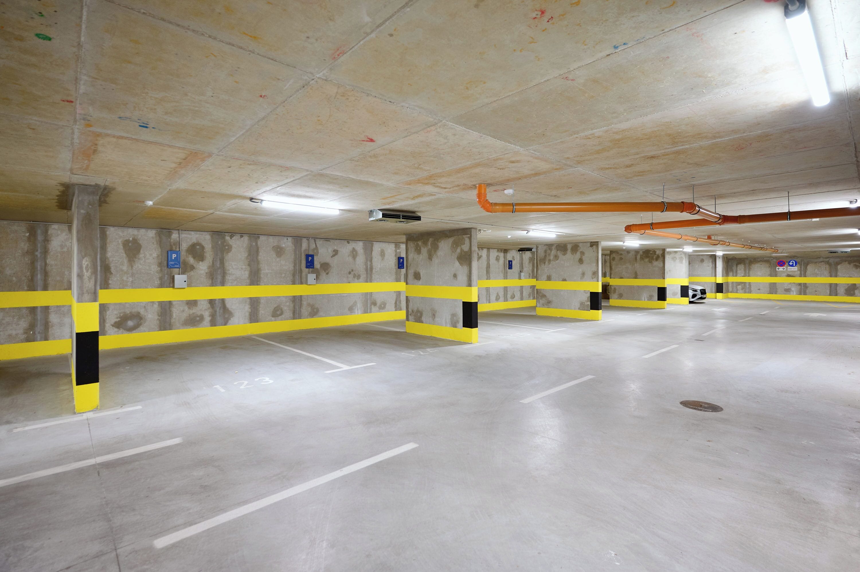 A multi-level parking garage with yellow and black safety barriers, concrete pillars, and overhead pipes.