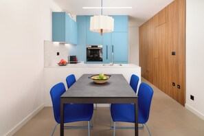 A modern kitchen with blue cabinets, a central island with a bowl of fruit, and a dining area with blue chairs.