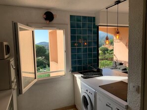 A compact kitchen with a washing machine, a window with an open door, and a view of greenery outside. - Ocean view & terrace – 10 min from Saint-Tropez – Pool, tennis court, private parking (La Croix-Valmer)