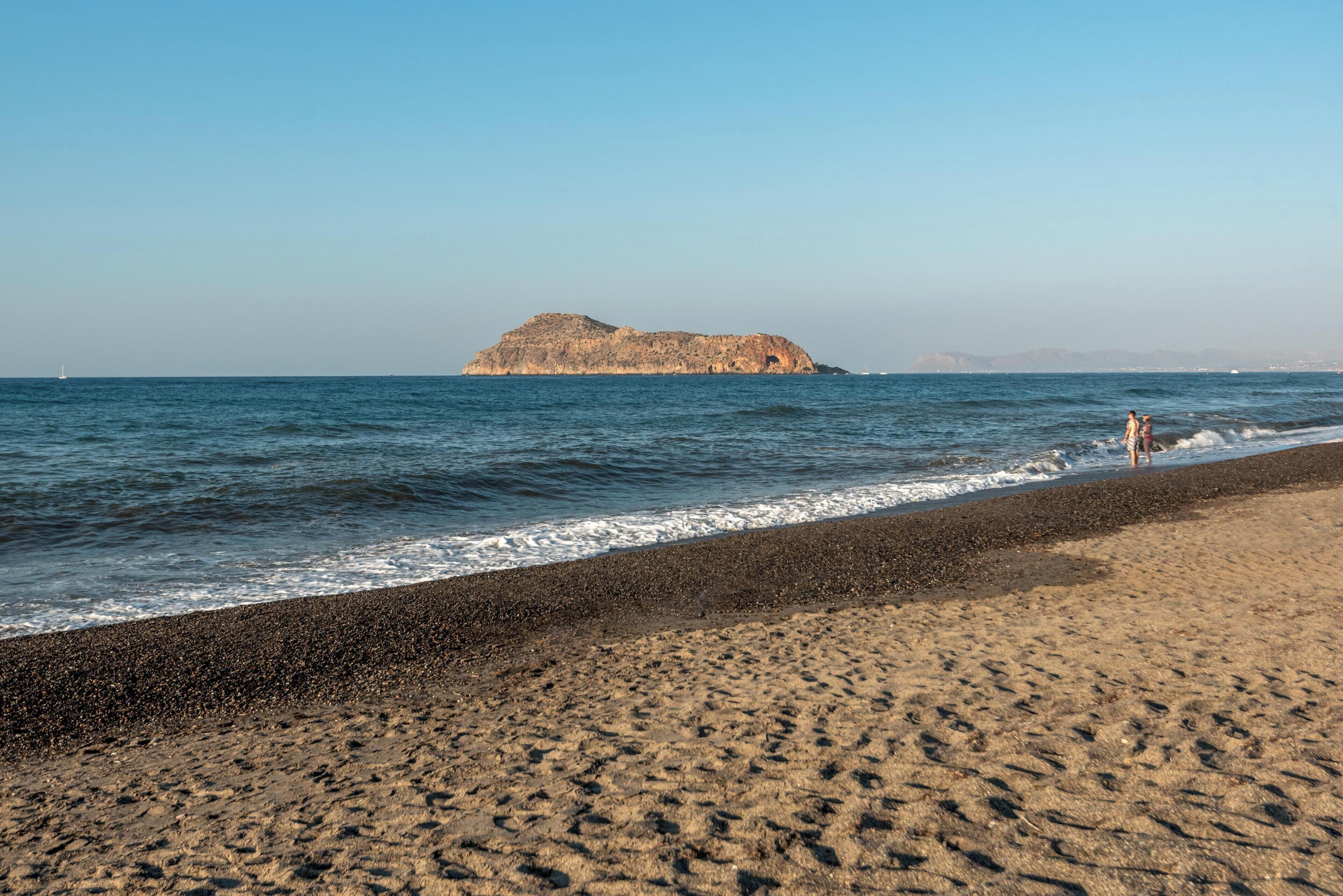 A beach with a rocky outcrop in the distance, a pebbly shore, and a clear sky.