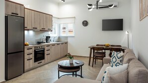 A modern kitchen with a refrigerator, oven, and coffee maker, a dining area with a table and chairs, and a living room with a sofa and a TV mounted on the wall. - Marilianas Apartment (Platanias)
